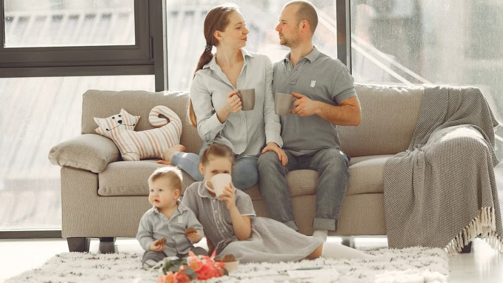 Happy family with children spending time at home while parents sitting on couch with cups of coffee and looking at each other when kids resting on floor